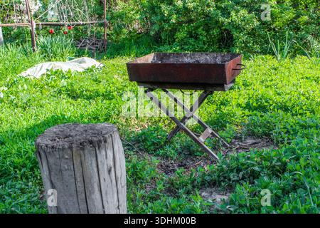 Brazier with ashes from firewood on the green grass background. Metal barbecue grill after cooking shashlik. Old small stump is used as a stool. Stock Photo