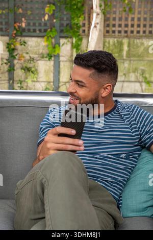 Adult male sitting on grey sofa wearing striped tee holding smartphone and looking left in backyard Stock Photo