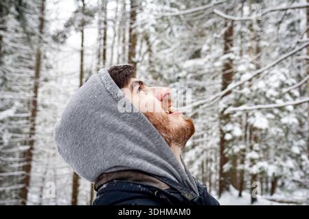 Bearded man in a hood joyfully looking up at falling snow, enjoying winter moment in a beautiful snowy forest. Authentic winter lifestyle, happiness Stock Photo