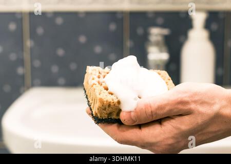 Focused shot of fingers gripping sponge laden with white suds, soap bottle and faucet blurred in background over white basin with blue walls. Stock Photo