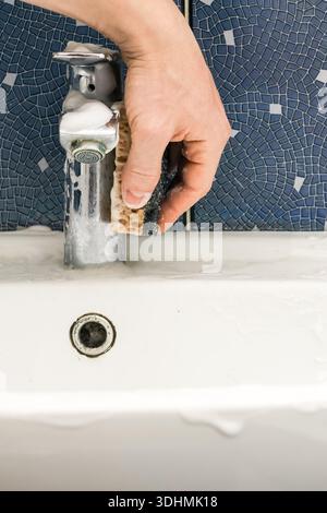 Detailed image of hand using foam-covered sponge on chrome tap in tiled sink area. Stock Photo