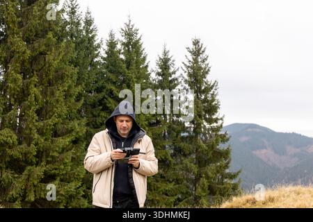 Man flying drone in mountain forest landscape. Middle-aged man in hooded jacket operating drone with remote controller in coniferous forest near Stock Photo