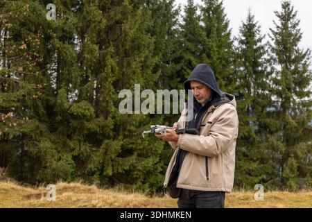 Man preparing drone for flight in forest. Adult male in hooded jacket setting up drone in coniferous forest, possibly for nature photography or aerial Stock Photo