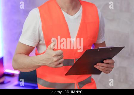 Confident male worker in orange safety vest showing approval gesture with clipboard in hand Stock Photo