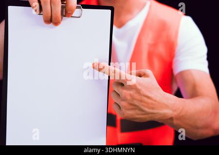 Male worker in fluorescent orange vest pointing finger at empty white sheet on clipboard against black backdrop Stock Photo