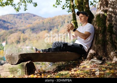 A focused man interacts with his smartphone, leaning back on a wooden bench beside an ancient mossy tree, surrounded by autumn leaves and a picturesqu Stock Photo