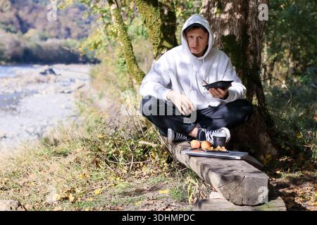 Young man in white hoodie sits cross-legged on log, holds black container, near apples, cup, cookies, tablet in forested area. Stock Photo