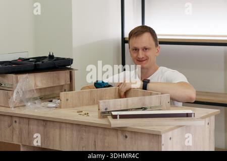 Smiling young man in white shirt sits at wooden table, holding blue drill near partially assembled light wood drawer with metal slides. Stock Photo
