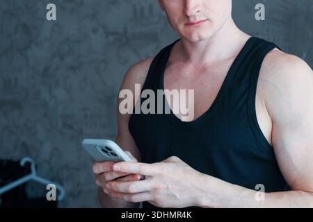 Athletic male in black sleeveless shirt holding mobile phone, focused gaze downward in indoor setting Stock Photo