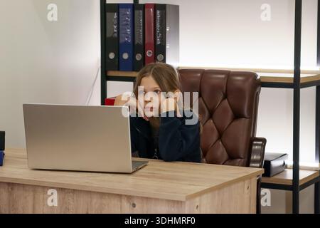 A preteen girl sits frustrated at a wooden desk, propping her head with hands, staring at a closed laptop amid shelves of binders. Stock Photo