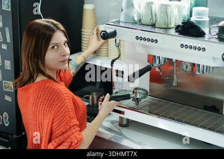 Back view of female barista in orange sweater working on espresso machine, holding portafilter and glancing back in cafe setting. Stock Photo