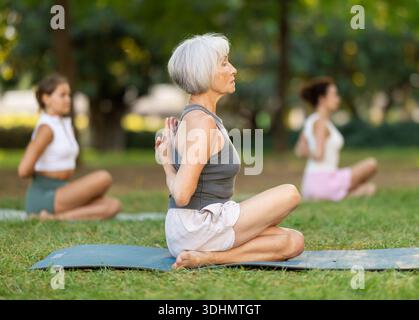 Elderly woman performing Reverse Prayer Gomukhasana during group yoga in park Stock Photo