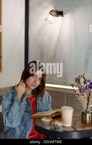 Young woman touching hair while reading book at cafe table with coffee, muffins and dried flowers Stock Photo