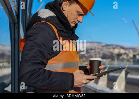 Profile of safety worker scrolling on smartphone, cup in other hand, clipboard under arm, against winter cityscape and blue sky. Stock Photo