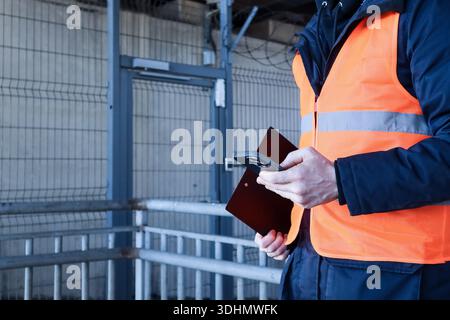Detailed view of gloved hands holding smartphone and black clipboard in orange vested worker at industrial setting Stock Photo