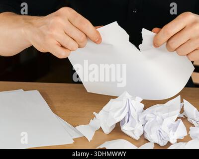 Close-up of person tearing white sheet in two amid crumpled and torn papers on wooden table Stock Photo