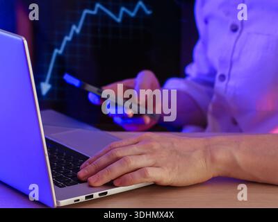 Close-up of hands on laptop and smartphone, downward trending financial chart visible in blue-toned background Stock Photo