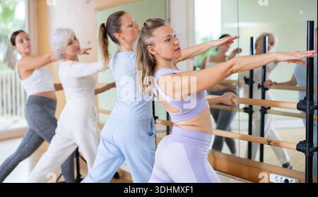 Concentrated women of different ages performing graceful hand movements near the barre, standing in a ballet pose during training in gym Stock Photo