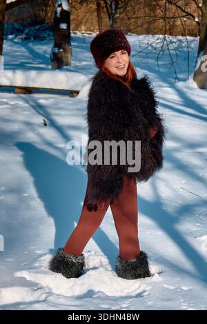 This stock photo showcases a beautiful, smiling woman with red hair enjoying a snowy winter day outdoors. She is wearing a stylish and warm fur hat, a Stock Photo