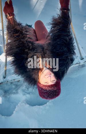This image captures a cheerful red-haired woman, clad in cozy winter attire, playfully suspended upside down on a swing. The backdrop showcases a sere Stock Photo