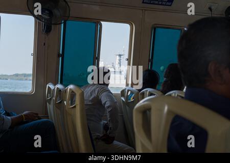 Kochi, India - February 27, 2023: Several passengers are seated inside a ferry, looking out through blue-tinted windows at a large ship passing on the water, with plastic seats and a wall-mounted fan visible in the cabin. Stock Photo