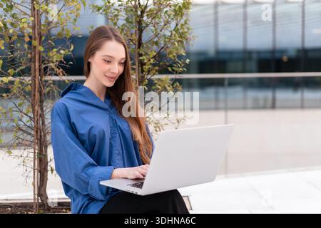 Confident female entrepreneur in blue shirt working on laptop against plants and office building. Beautiful businesswoman in smart casual using netbook during daytime Stock Photo