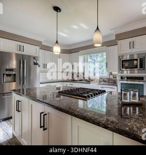 Bright modern kitchen with white cabinetry, stone countertops, stainless steel appliances, gas cooktop island, pendant lighting, and natural daylight. Stock Photo