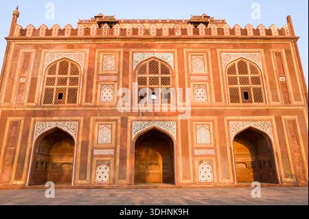 Agra, India - March 12, 2024: The image shows the symmetrical facade of the west gate at Akbar's Tomb, featuring large arched entrances, geometric stone latticework, carved red sandstone panels, and decorative white inlay illuminated by sunlight. Stock Photo