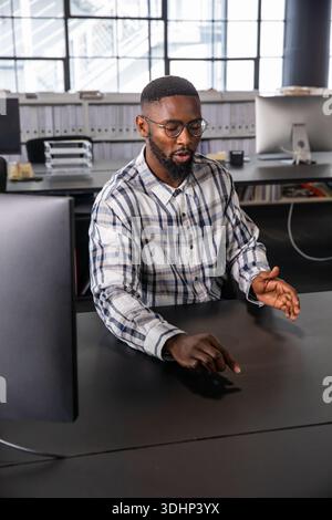 Mid-adult African American man gesturing, pointing at desk in office with monitor, binders, glasses Stock Photo