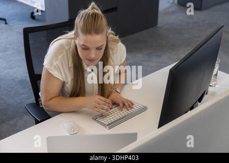 Female worker leaning over typing on slim keyboard at white desk in office with laptop, monitor Stock Photo