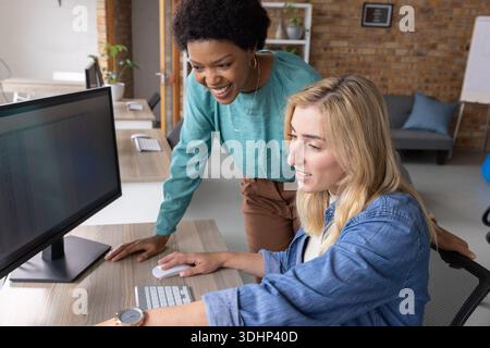 Diverse female coworkers collaborating at desk in open-plan office, viewing spreadsheet on monitor Stock Photo