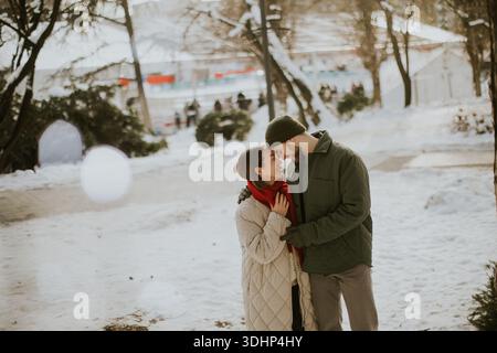 A Korean woman and a Caucasian man are standing close in a winter park. They share a warm moment surrounded by snow and trees. People walk in the back Stock Photo