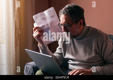 Stressed middle aged man sitting at home reading bills and paperwork on a laptop, dealing with financial pressure, responsibility and mental overload. Stock Photo