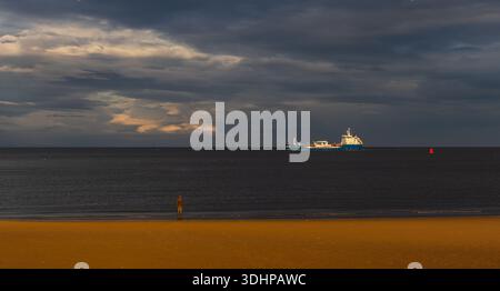 Figure from Antony Gormley’s Another Place installation on the beach at Crosby with a cargo ship offshore Stock Photo