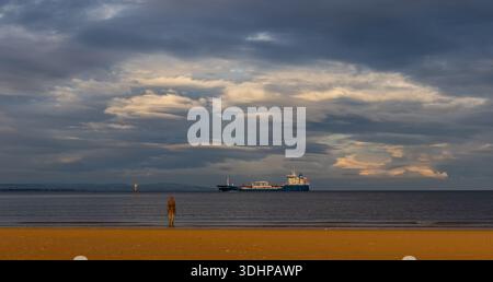 Figure from Antony Gormley’s Another Place installation on the beach at Crosby with a cargo ship offshore Stock Photo