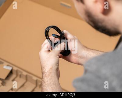 Close-up of a man's hands holding a coiled black USB data transfer cable from a cardboard box, emphasizing the process of connecting and configuring n Stock Photo