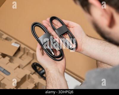 Close-up of a person holding two new black USB data and charging cables above an opened brown cardboard box, highlighting technology unboxing and acce Stock Photo