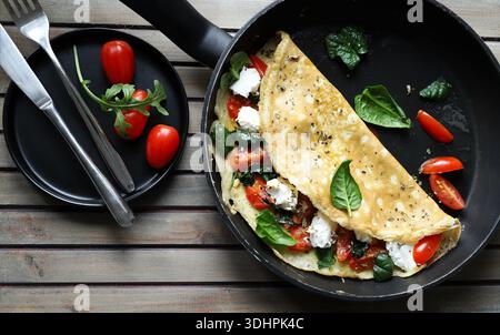Savory pan-fried omelette filled with fresh spinach, cherry tomatoes, and cheese on a wooden background. Overhead view. Stock Photo