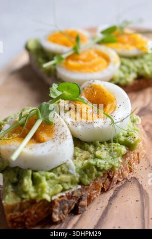 Close‑up photograph of avocado toast topped with soft‑boiled eggs, black pepper and fresh sprouts, served on a wooden board with cutlery. The warm ton Stock Photo