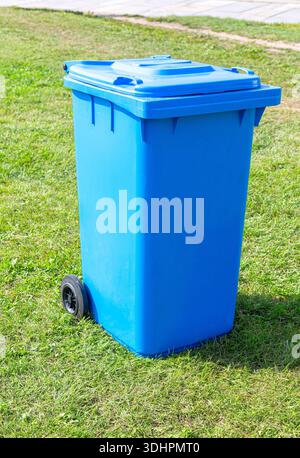 Covered blue plastic garbage container with wheels on the green grass in summer Stock Photo