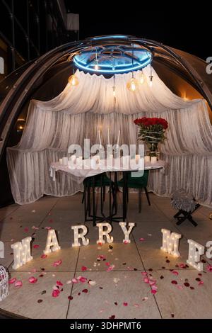 A beautifully arranged romantic proposal scene under a glass dome with white drapery, candles, red roses, and illuminated "MARRY ME" letters surrounde Stock Photo