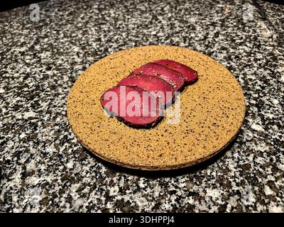 Four thin slices of rare, seasoned meat are artfully arranged on a circular, textured serving plate, set on a speckled granite surface. Stock Photo