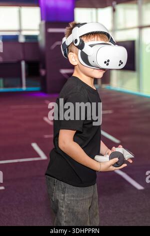 Child wearing VR glasses holds a controller while exploring a virtual space in a gaming center during daytime Stock Photo