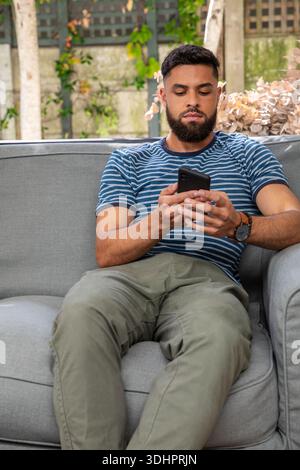 Bearded man sitting on gray sofa in patio, wearing striped T shirt and watch, holding smartphone Stock Photo