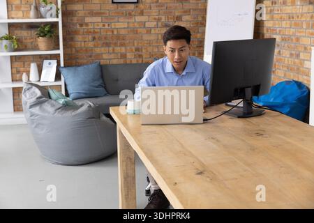 Asian man working on laptop with external monitor at home office desk, wearing light blue shirt Stock Photo