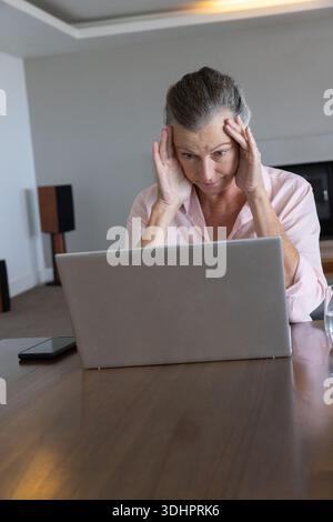 Senior woman rubbing temples, wearing light pink shirt at home, working on laptop with phone, glass Stock Photo