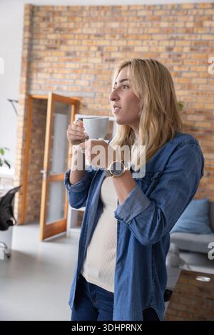 Mid-adult woman standing, holding white ceramic cup and saucer, looking right in loft workspace Stock Photo