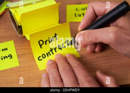 from above, a yellow sticky note reading print contract is held steady while a man finishes writing with a black marker on a wooden office desk Stock Photo