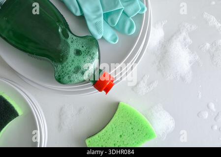 Hand washing dishes background with stack of dishes and detergent gloves and scouring pads on top on a white kitchen bench with water and foam. Top vi Stock Photo