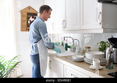 Detail of man with gloves and apron washing dishes in a white kitchen. Side view. Stock Photo
